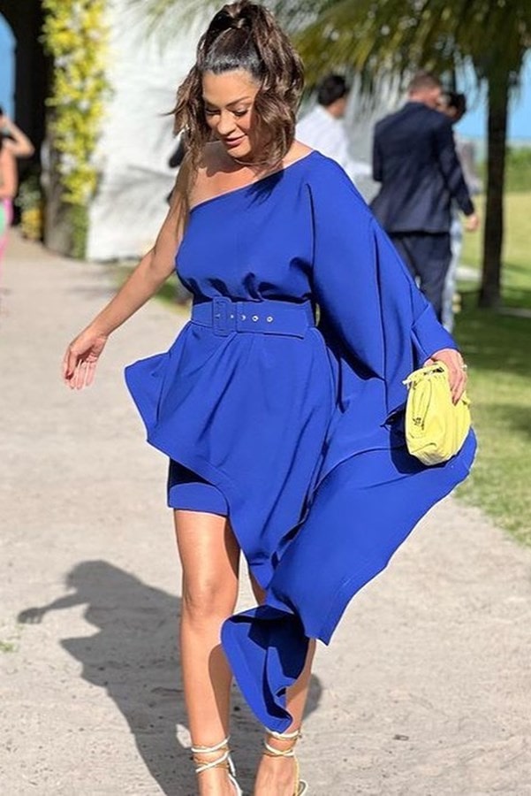 convidada de casamento na praia usando vestido curto azul modelo um ombro só com uma manga capa e cinto do tecido do vestido. Ela usa ponytail e nas mãos segura uma clutch amarelo lima